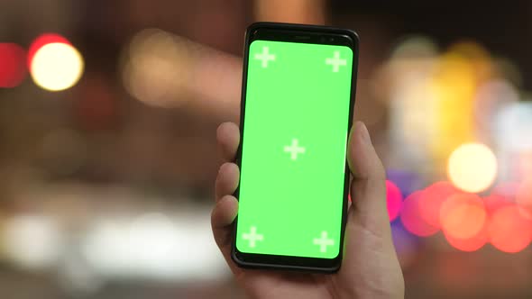 Closeup of Young Man Showing Phone with Green Screen Against Illuminated Lights at Night