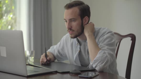 Confident Bearded Man Draws Using Graphic Tablet Looking Into a Laptop