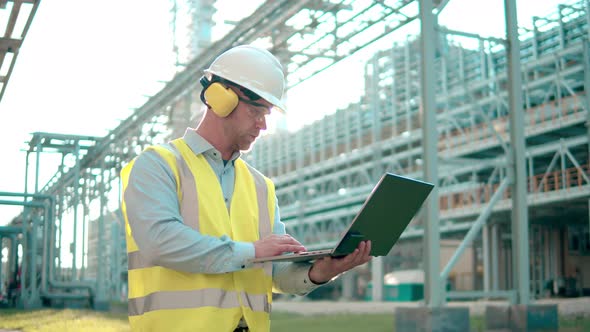 Refinery Engineer in a Hardhat and Headphones is Operating a Laptop at a Modern Industrial Facility