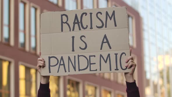 Male Hands Hold a Poster with the Slogan RACISM IS A PANDEMIC Against the Background of the City