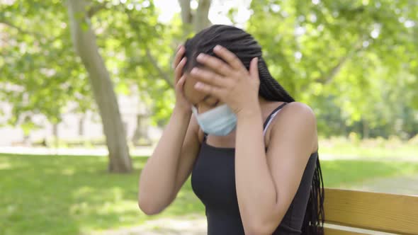 A Young Black Woman in a Face Mask Acts Upset and Sits on a Bench in a Park on a Sunny Day