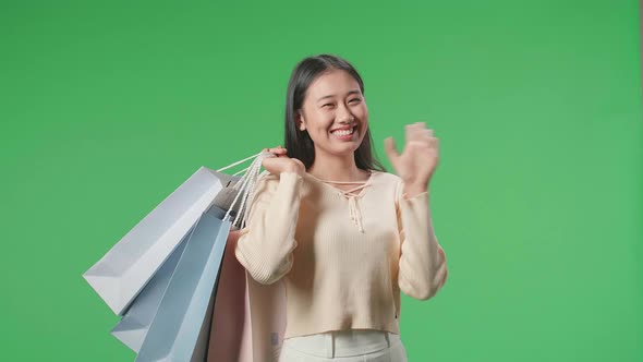 Asian Shopping Woman Holding Shopping Bags Up Before Waving Hands And Smiling At Green Screen Studio