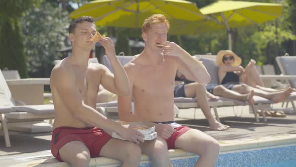 Two Young Caucasian Men Sitting at Poolside and Eating Tasty Pizza. Portrait of Happy Relaxed Male