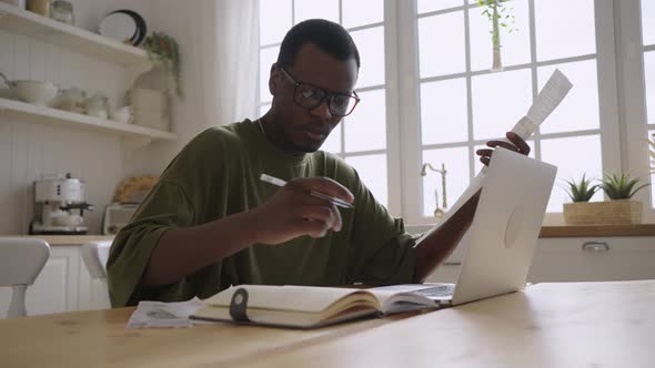 African American Man Looks at Checks Near Laptop in Kitchen