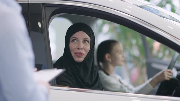 Gorgeous Slim Woman in Hijab Talking To Unrecognizable Dealer with Blurred Girl Sitting on Driver's