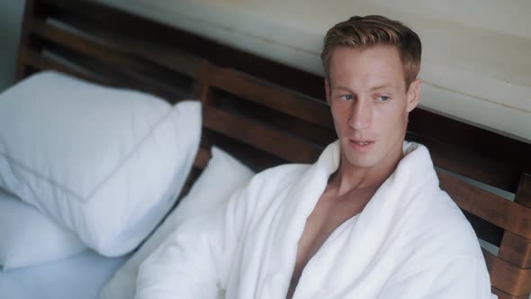 Handsome Man in White Bathrobe Lies on Bed in His Apartment and Drinks Coffee