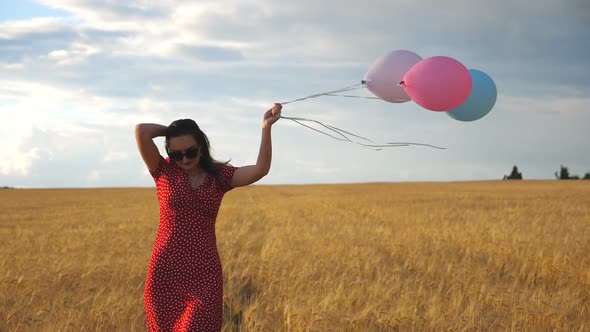 Happy Beautiful Girl in Sunglasses Walking Through Wheat Field and Holding Balloons in Hand