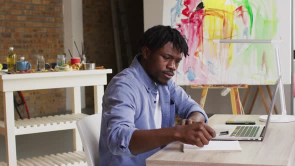 African american male artist taking notes while sitting on his desk at art studio