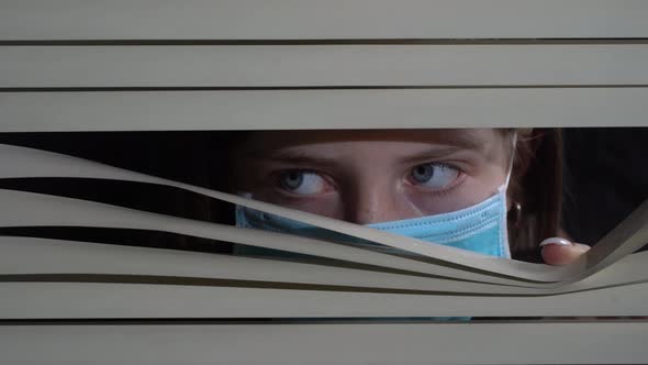 Girl in protective mask looking out the window through the blinds to street