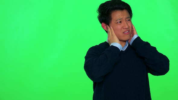 A Young Asian Man Covers His Ears and Grimaces Against a Loud Sound - Green Screen Studio