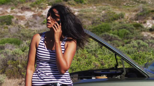 African american woman talking on smartphone while standing near her broken down car on road