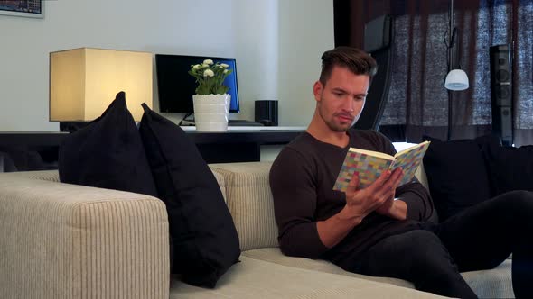 A Young, Handsome Man Reads a Book on a Couch in a Cozy Living Room