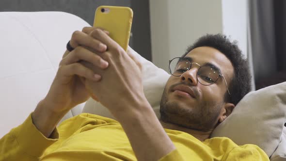 African American Man Lies Resting on the Couch at Home and Holds a Smartphone