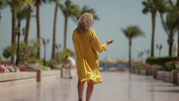 a Curly Blonde in Sunglasses and a Yellow Fluttering Dress Looks at the Phone and Walks Away Against