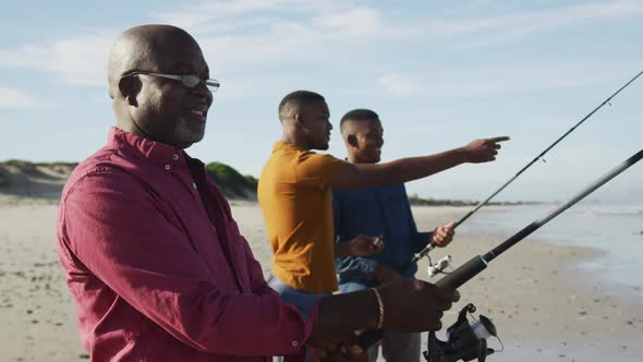 African american senior father and twin teenage sons standing on a beach fishing and talking