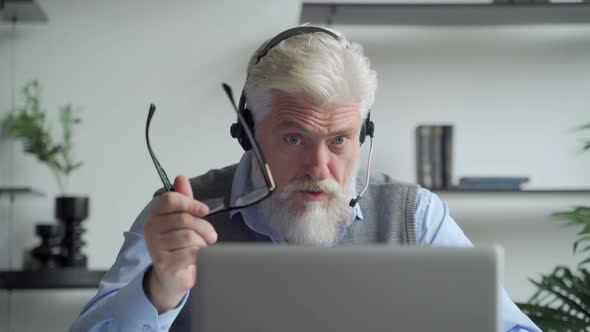 Smiling Elderly Man with a Gray Beard Wearing Headphones, Having a Video Call on a Laptop, Happy