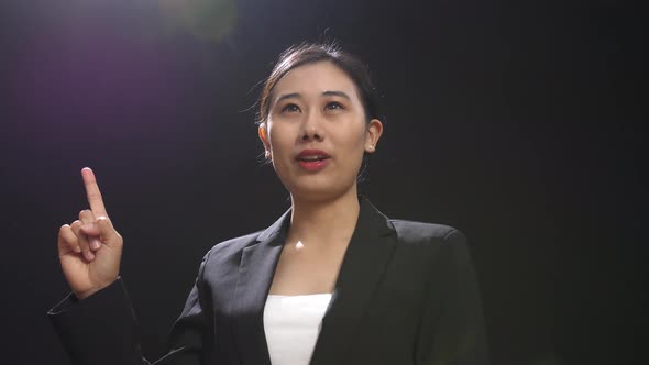 Asian Speaker Woman In Business Suit Showing Index Fingers Up While Speaking In The Black Studio