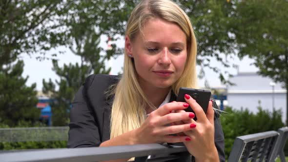 A Young Beautiful Businesswoman Sits on a Bench and Works on a Smartphone in a Park - Closeup