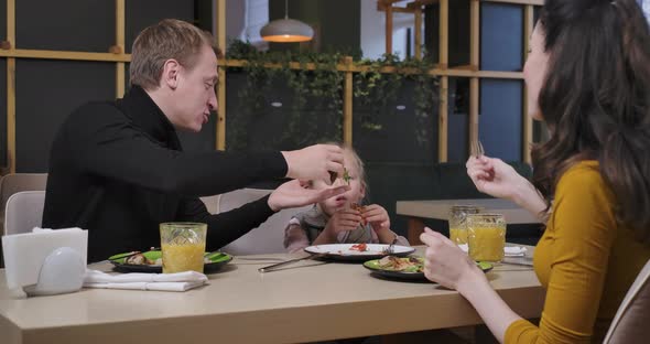 Happy Cute Girl Eating Pizza with Parents in Restaurant Indoors