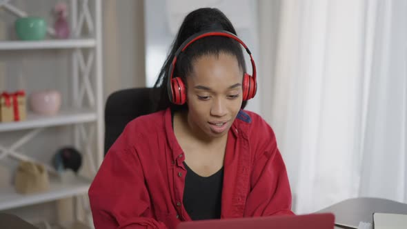 Cheerful Female Freelancer Dancing to Music in Headphones Sitting at Table Typing on Laptop Keyboard