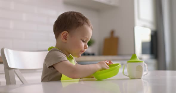 Cute Little Baby Boy Sits at a Table in the Kitchen and Eats Strawberries From Green Plate with His