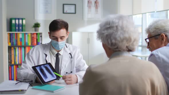 Young Doctor in Safety Mask Show Xray on Tablet to Aged Couple