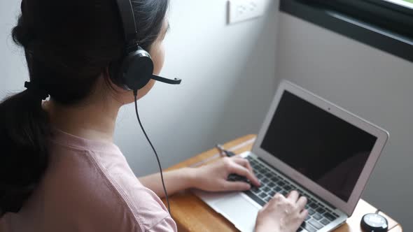 unrecognizable Asian woman wearing headset, using laptop computer near windows, working, talking.