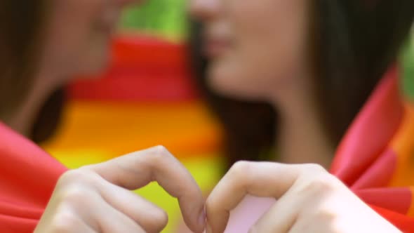 Beautiful Lesbians Wrapping in Rainbow Flag, Making Heart With Hands and Kissing