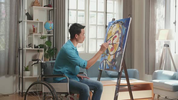 Side View Of An Asian Artist Man In Wheelchair And Painting A Girl On The Canvas