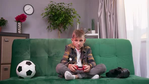 Modern Teen Boy Sitting on Comfortable Sofa at Home and Applying Joystick while Playing Video Game