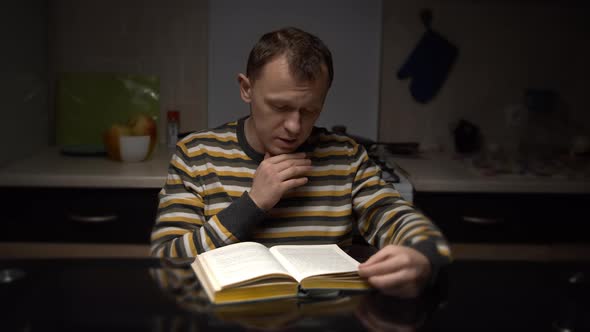 Young Man with Expression Reads Text From a Book While Sitting in the Evening at the Table