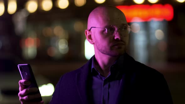 A Bald Solid Guy in a Blue Jacket Uses a Mobile Phone on the Street at Night