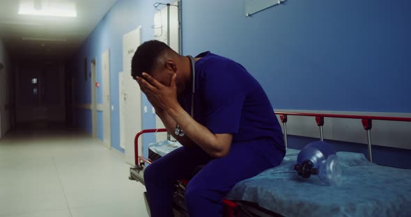 A Doctor Sits on a Medical Gurney and Rubs His Head in Empty Hospital Corridor