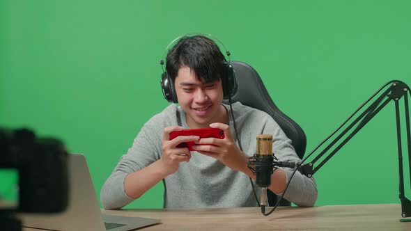 Smiling Asian Man Gamer With Headphone Looking At Camera While Using Mobile Phone On Green Screen