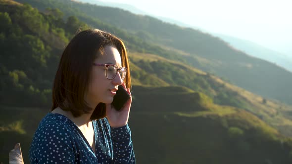 A Young Woman in a Dress Sits and Speaks on the Phone Against a Background of Mountains. The Girl