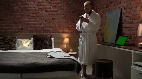 A handsome young businessman stands in a bathrobe after a shower with a phone in his hands