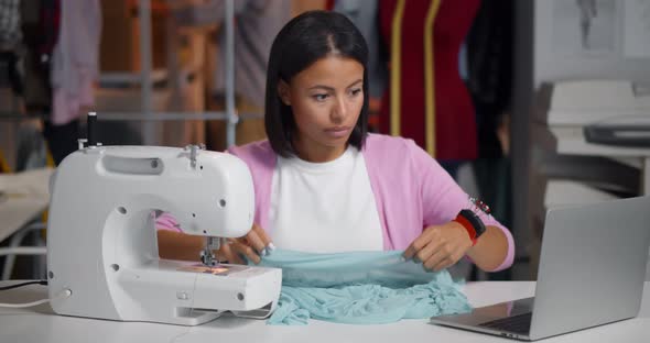 African Female Tailor Working with Fabric and Using Laptop Sitting at Desk in Workshop