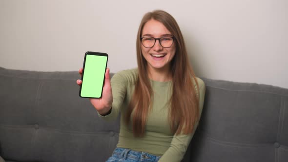 Pretty Laughs Woman in Glasses Showing Smartphone with Green Screen Chromakey on Couch at Home