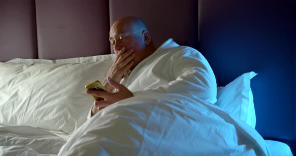 Man Is Lying in Bed. He Holds a Smartphone in His Hand, but Wants To Sleep, Yawns, Rubs His Eyes