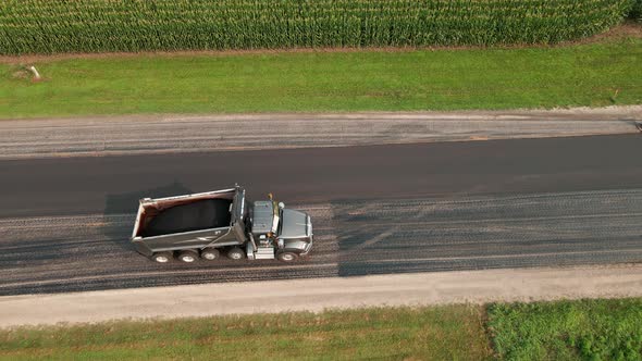 Birds eye view of dump truck backing down the highway with aggregate to fill paver on rural road.