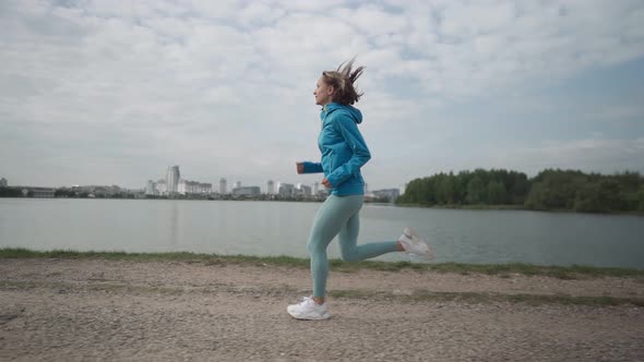 Slow Motion Young Sportswoman in Sportswear Trains in a Park Near the River