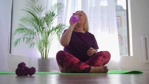 Fitness Training  Blonde Overweight Woman Sitting on Yoga Mat and Drinking Water From the Bottle