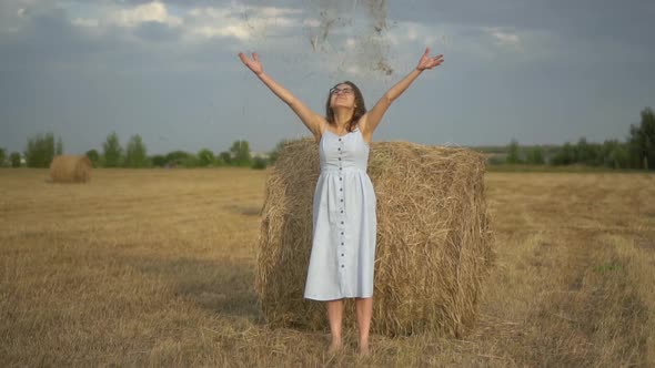 Young Woman Throws Dry Grass While Standing By a Haystack in a Field