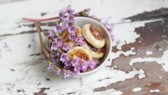 A Bouquet of Lilac Flowers Near a Plate with Cheese Cakes on a Wooden Background