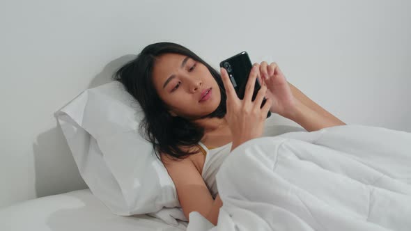 Young Asian woman using smartphone checking social media feeling happy smiling while lying on bed.