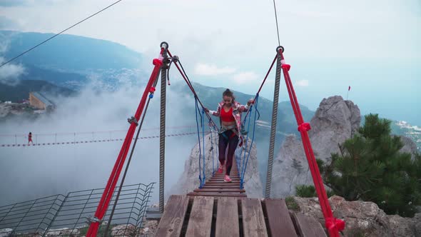 Young Athletic Girl Is a Traveler Crossing the Rope Bridge Above the Precipice in the Mountains
