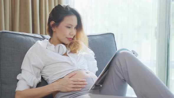 Asian young woman pregnant sitting and reading book with smile on sofa.