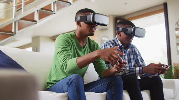 African american teenage twin brothers sitting on couch using vr headsets and playing computer game
