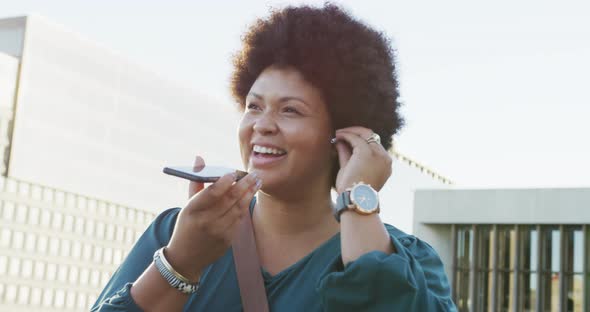 Happy plus size biracial woman talking on smartphone and wearing earphones in city