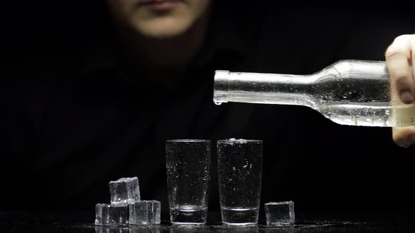 Barman Pour Frozen Vodka From Bottle Into Two Glasses with Ice. Black Background